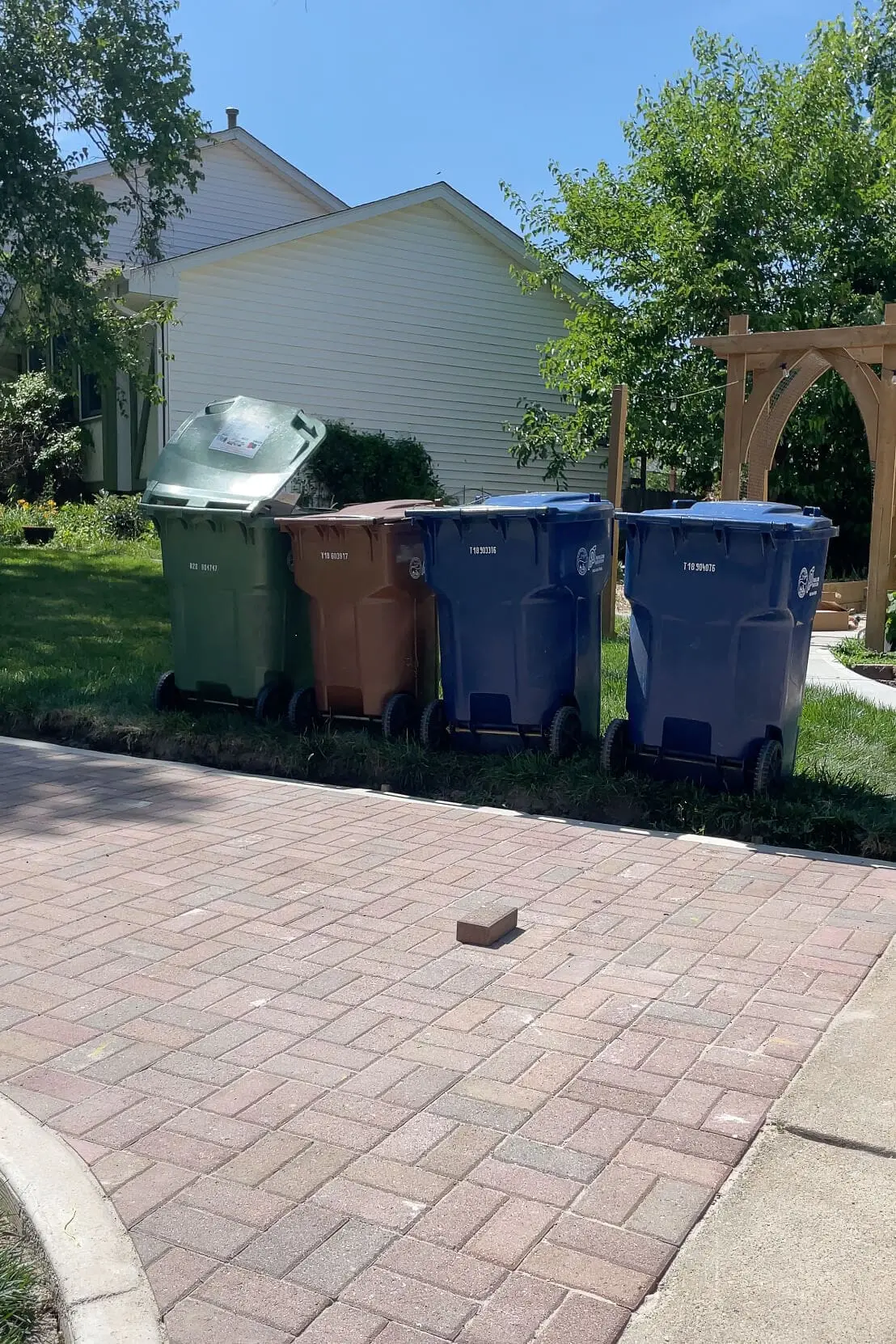 Four city-issued garbage and recycling bins lined up on the grass next to a paver driveway, awaiting a new enclosure.